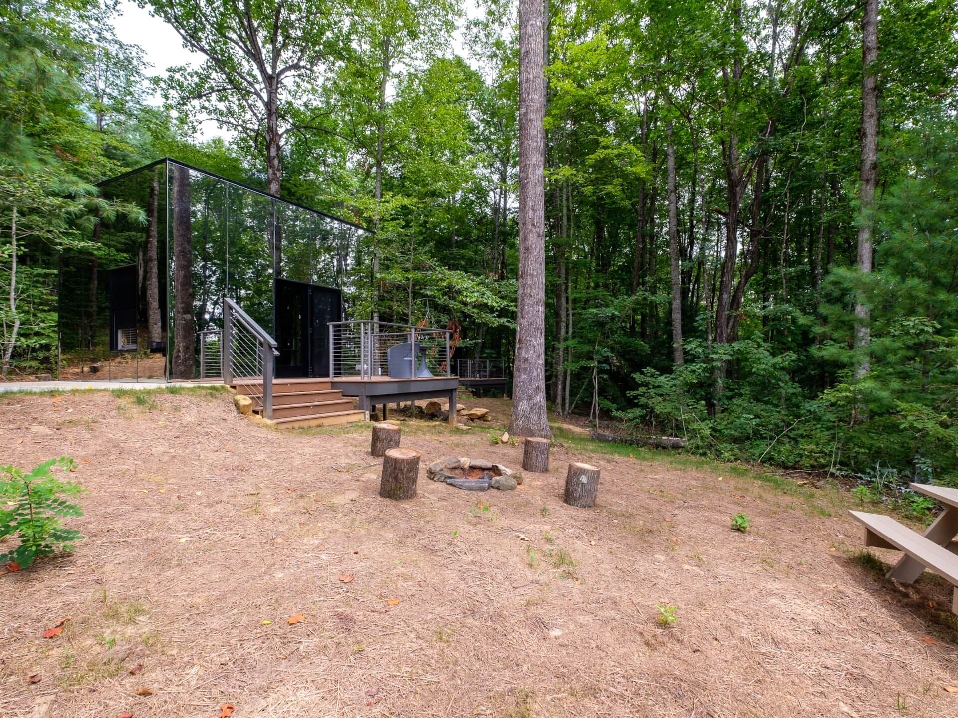 A modern outdoor structure with a glass enclosure, wooden steps, and a picnic table in a forested area.