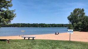 A beach with a lifeguard sign, a bench, and a group of people sitting on the sand. - Rome Ranch Retreat: Golf + Lake + Pool + ATV + Dog + Disc & Foot Golf too! (Nekoosa)