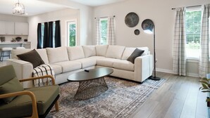 Een moderne woonkamer met een beige modulaire bank, een ronde salontafel en een vloerlamp. Er is een keuken met witte kasten en een eettafel.