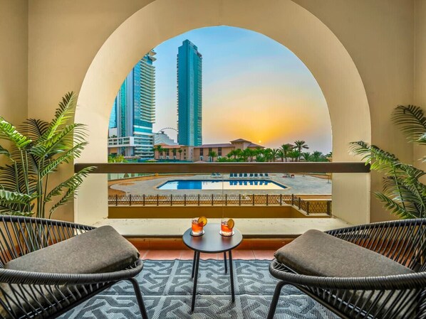 A balcony with wicker chairs, a small table, and a view of a cityscape with tall buildings and a pool.