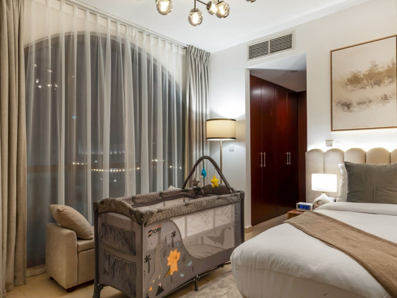 A hotel room with a large bed, a side table with a lamp, a baby playpen, a window with curtains, and a door leading to another room.