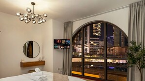 Quarto de hotel com uma cama grande, televisão na parede, uma janela ampla com vista para a cidade à noite e um vaso de plantas.