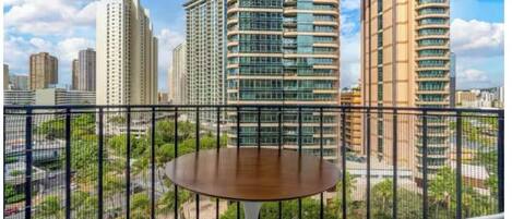 Uma varanda com uma mesa redonda e duas cadeiras, vista para um horizonte urbano com edifícios altos e áreas verdes.