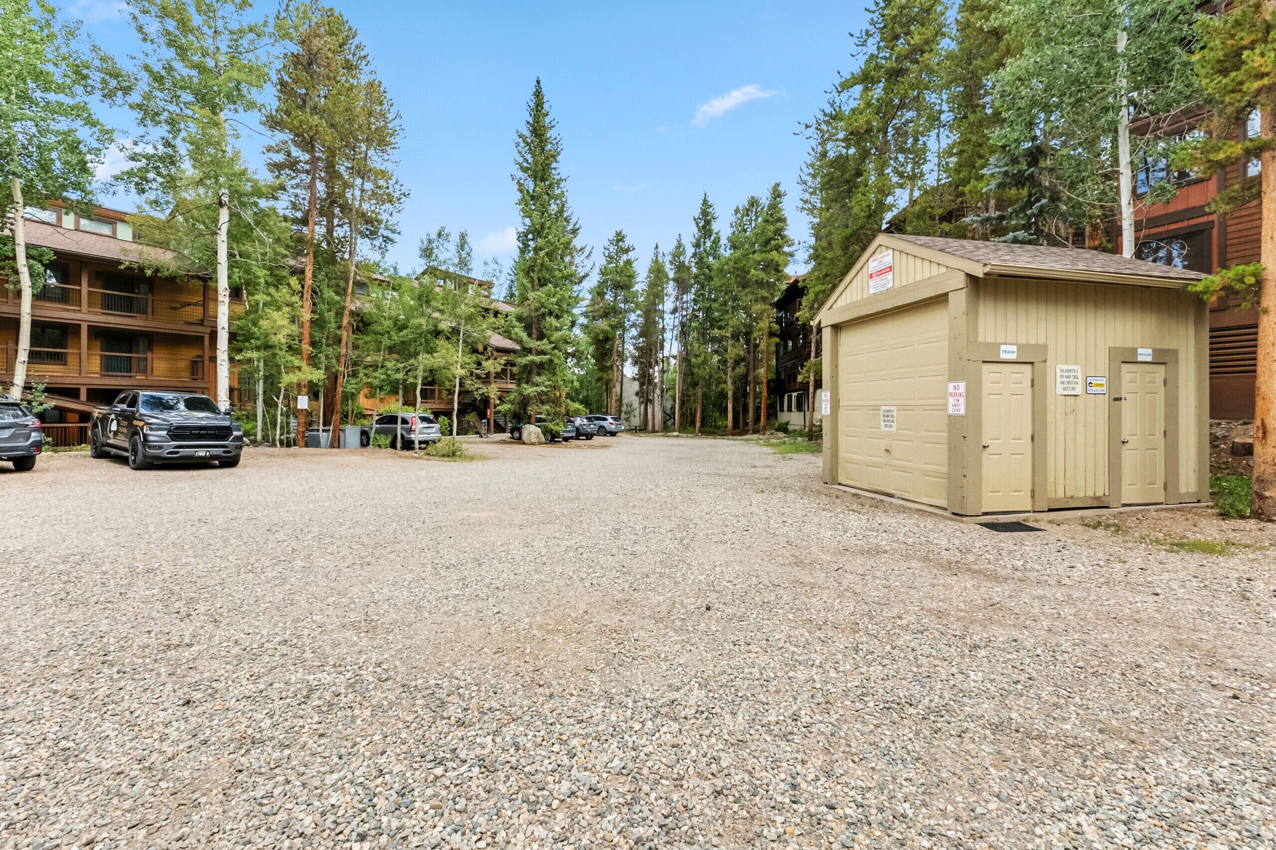 A gravel parking area with several cars, a wooden building with balconies, and a small shed with a sign.