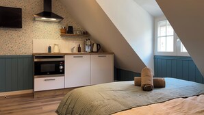A bedroom with a sloped ceiling, a bed with a green blanket, a wooden floor, and a kitchenette with a stove and cabinets. - English cottage - Atypical accommodation (Sarreguemines)