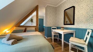 Ein gemütliches Schlafzimmer im Dachgeschoss mit schrägen Wänden, einem Bett mit zusammengerolltem Bettbezug, einem kleinen Tisch und einem Stuhl.