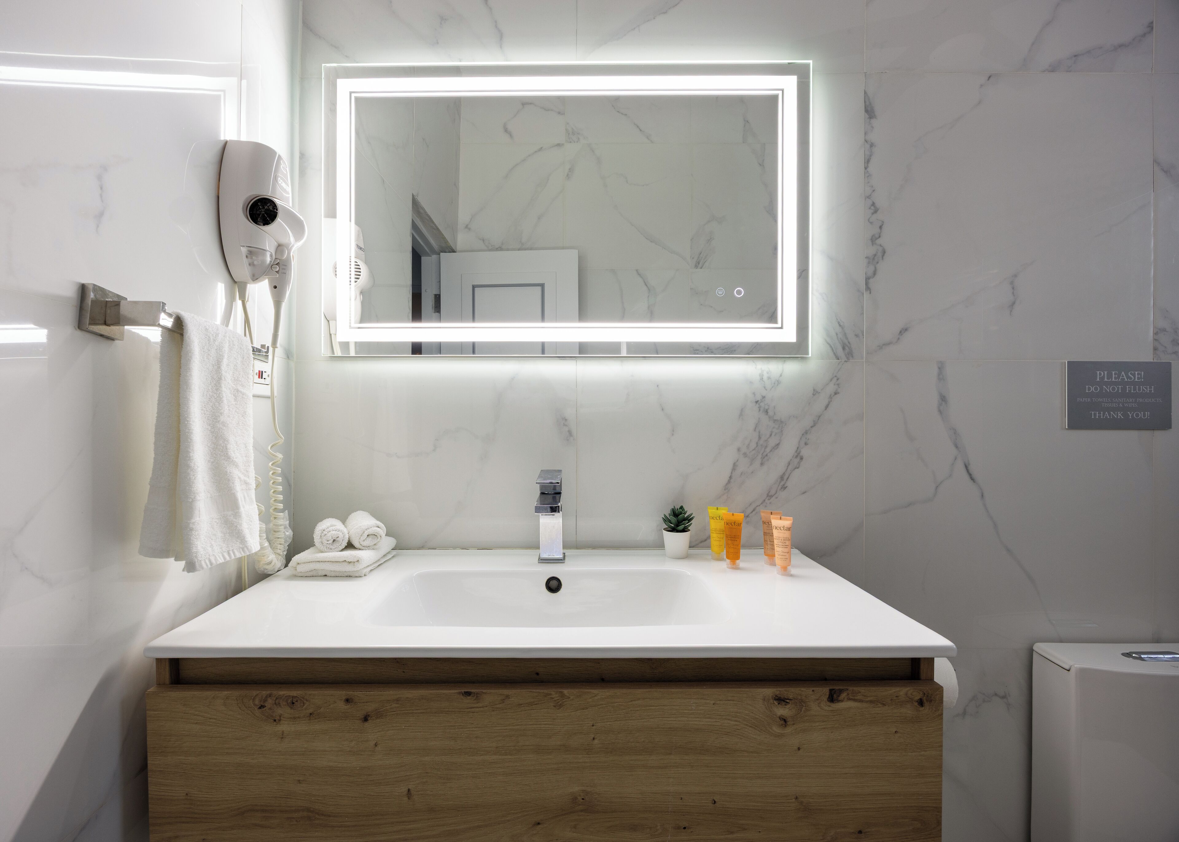 A modern bathroom with a marble backsplash, a large mirror with integrated lighting, a white sink, and a wooden vanity.