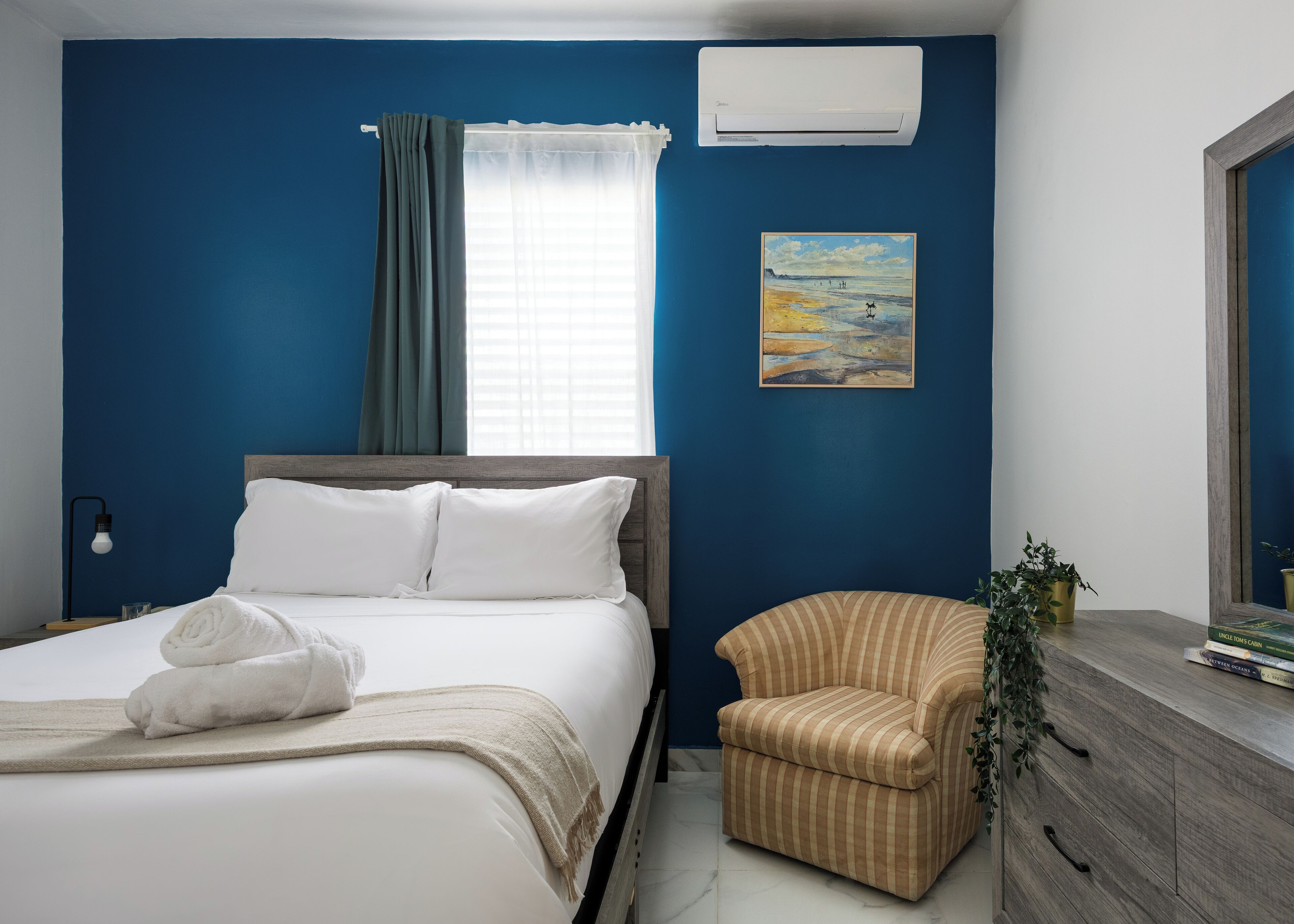 A bedroom with a bed, a chair, a dresser, a painting, and an air conditioner.