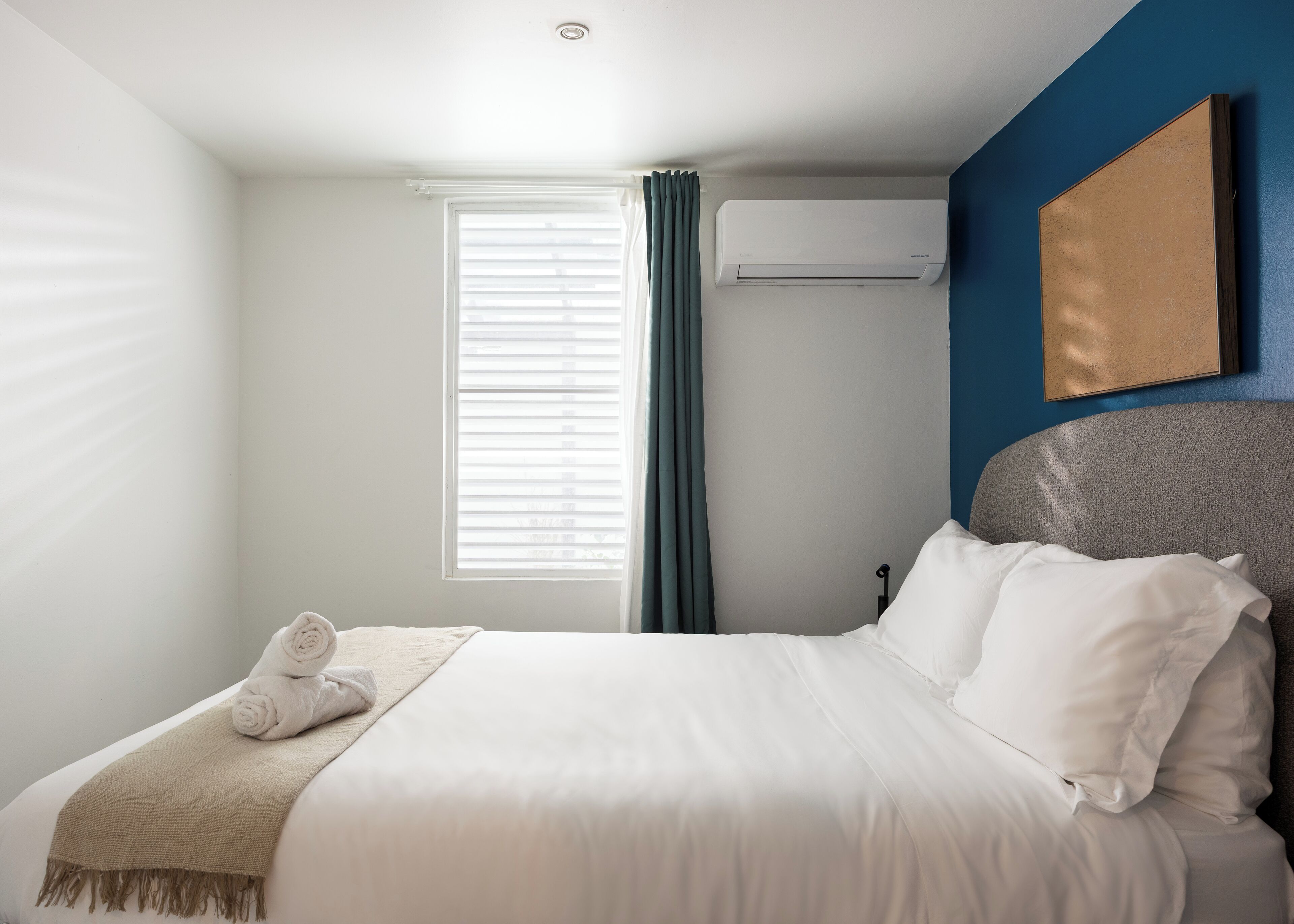 A neatly made bed with white linens and a beige throw blanket, a window with blinds, and an air conditioning unit on the wall.
