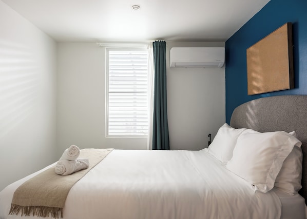 A neatly made bed with white linens and a beige throw blanket, a window with blinds, and an air conditioning unit on the wall. - Ocean Park 4BDR / 2b Pool @ Riviera Villa (San Juan)