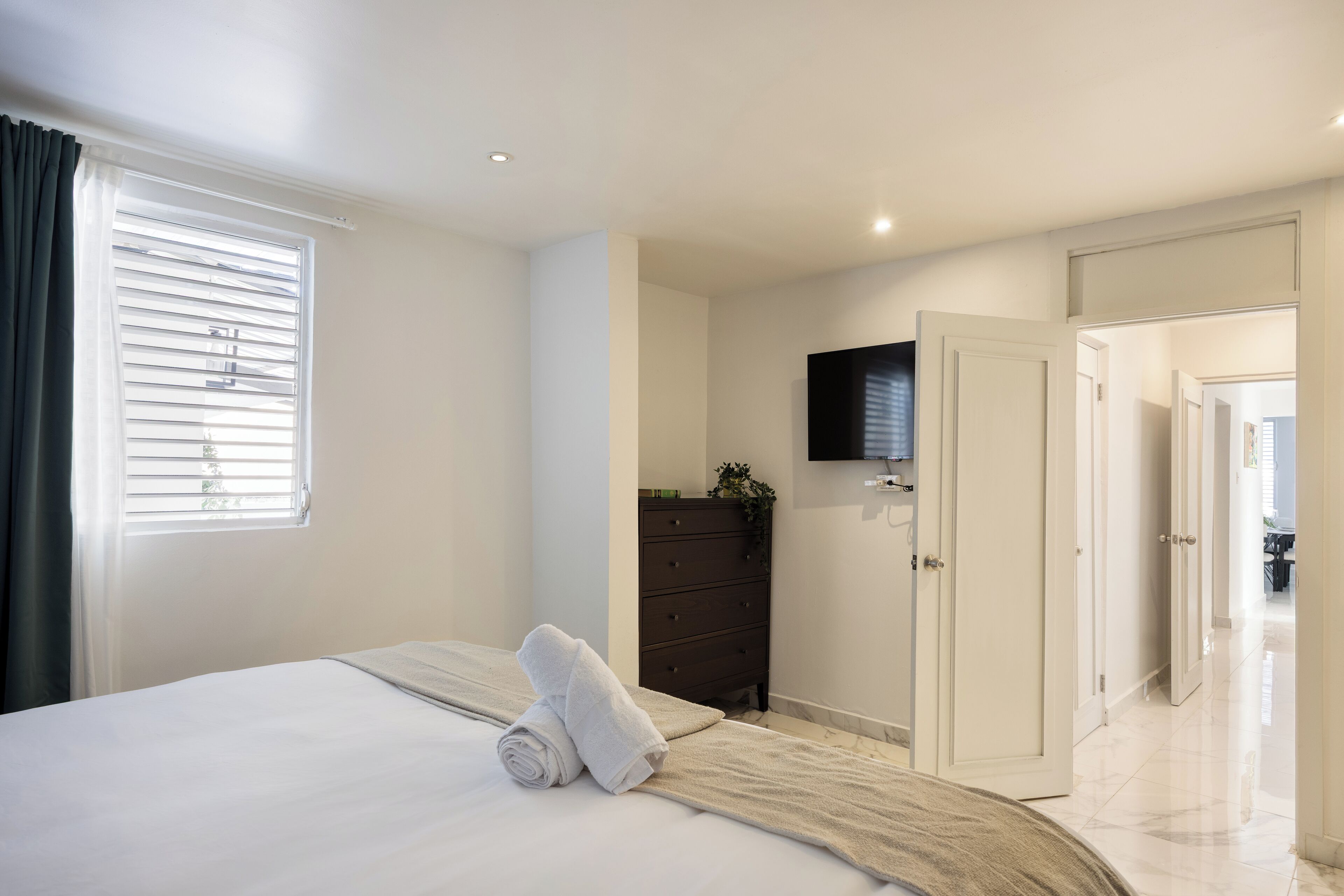 Une chambre à coucher comprenant un lit, une commode, un téléviseur fixé au mur et une porte donnant sur une autre pièce.
