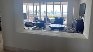 Una habitación con un ventanal grande que da a una piscina y al océano. Hay varias sillas y una mesa dispuestas cerca de la ventana.