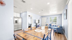 A modern kitchen with a stainless steel refrigerator, a dining table with chairs, and a living area with a sofa and a TV.