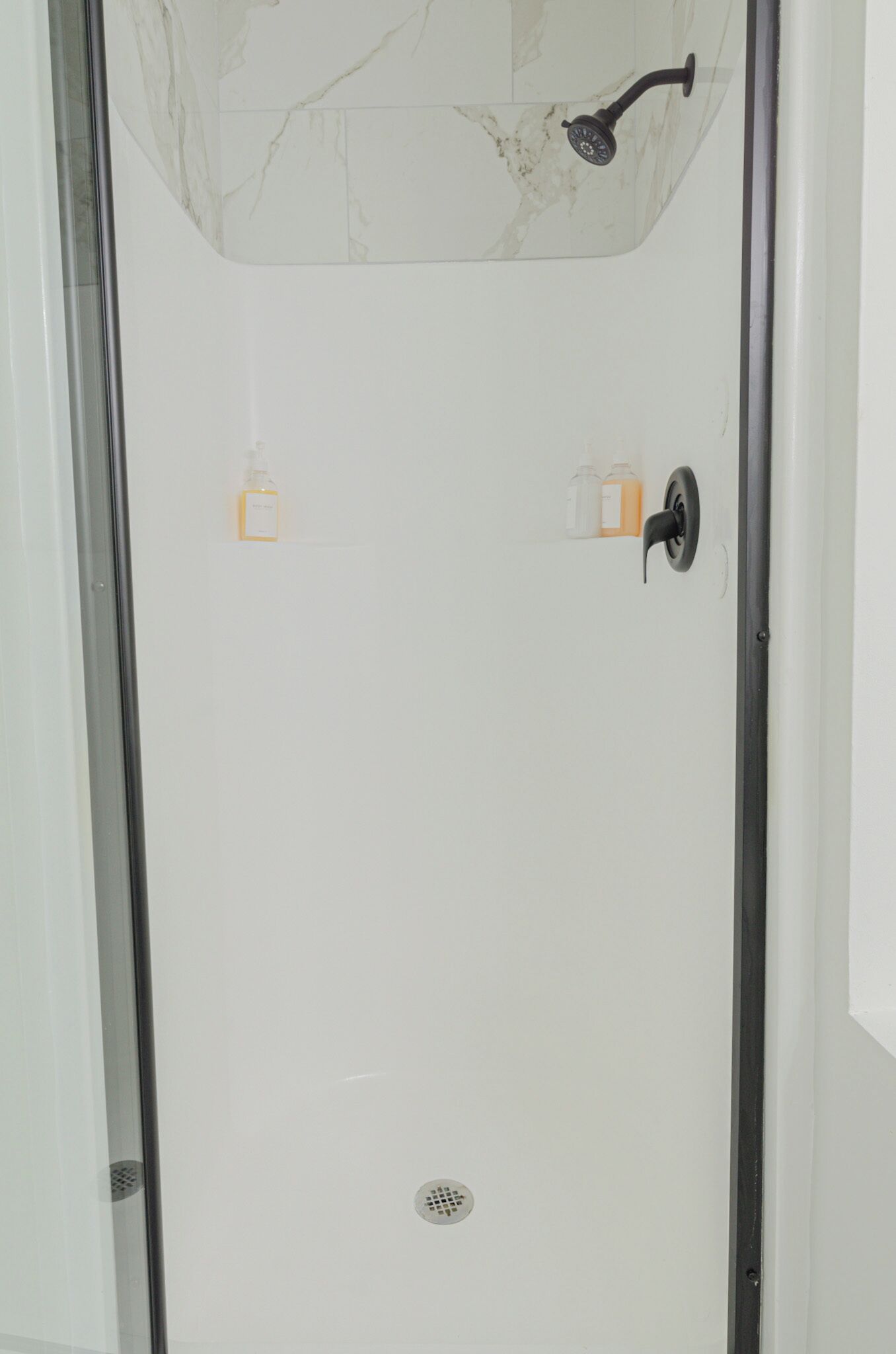 A glass-enclosed shower with a marble wall, a black showerhead, and two soap dispensers.