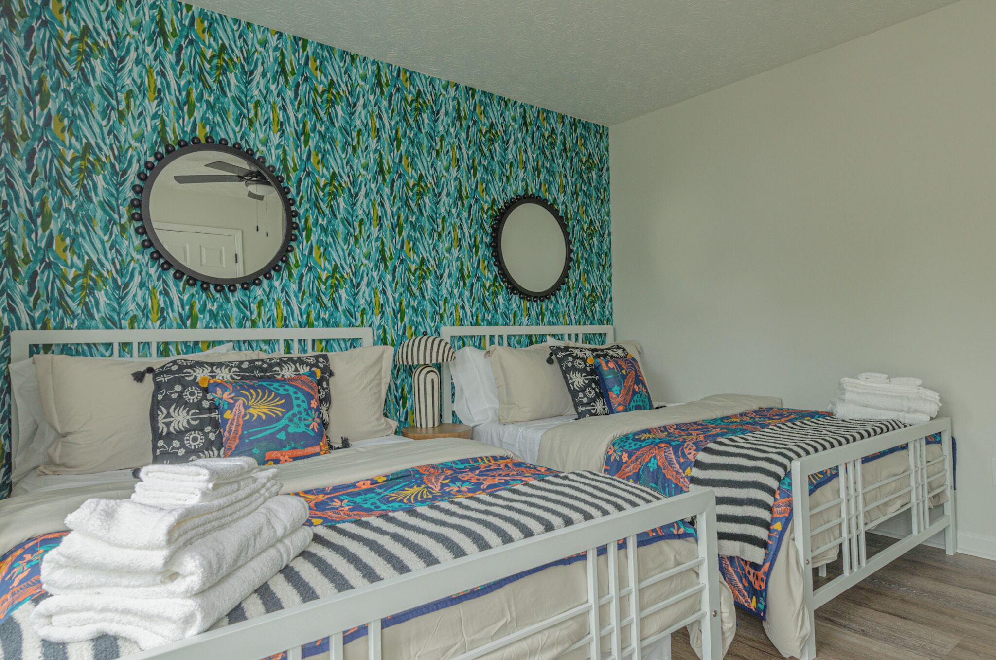 A bedroom with two beds, each with a white frame and patterned bedding. There are two round mirrors on the wall, and the room has a blue and green leafy wallpaper.