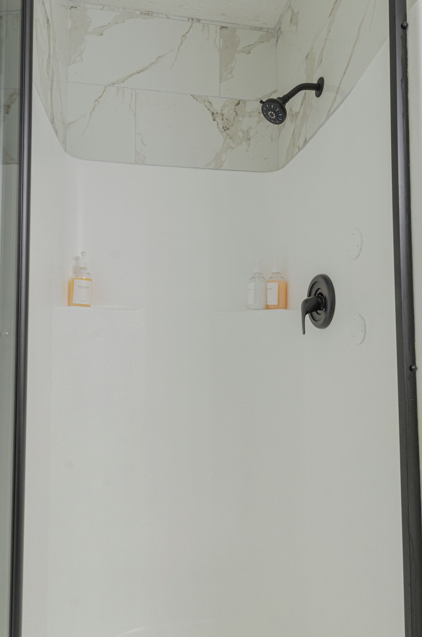 A bathroom with a marble shower enclosure, a handheld showerhead, and two bottles of liquid soap on the wall.