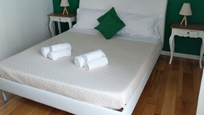 Una cama bien hecha con ropa blanca, dos toallas enrolladas, un cojín verde y dos mesitas de noche con lámparas.