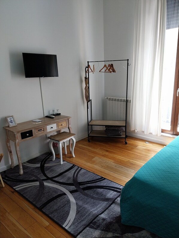 Une chambre avec un bureau en bois, un téléviseur fixé au mur, un lit recouvert d’une couverture turquoise, un radiateur et un porte-vêtements.