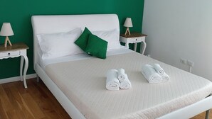 Una cama impecablemente hecha con sábanas blancas y dos toallas enrolladas, una almohada verde y un cabecero blanco. Hay dos mesitas de noche con lámparas, un tocador de madera y un aire acondicionado de pared.
