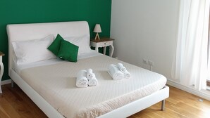 Una cama bien tendida con ropa blanca y almohadas verdes, una mesita de noche con una lámpara y una ventana con cortinas blancas.