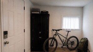 Une pièce avec une porte fermée, une armoire noire, un vélo, une fenêtre avec des stores et un sac à dos posé sur le sol.