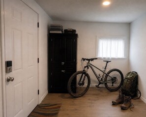 Una habitación con puerta cerrada, un armario negro, una bicicleta, una ventana con persianas y una mochila en el suelo.