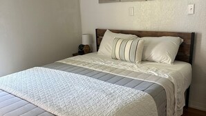 Ein ordentlich bezogenes Bett mit gesteppter Bettdecke und Kissen, ein Holz-Kopfteil und ein Nachttisch mit Lampe.