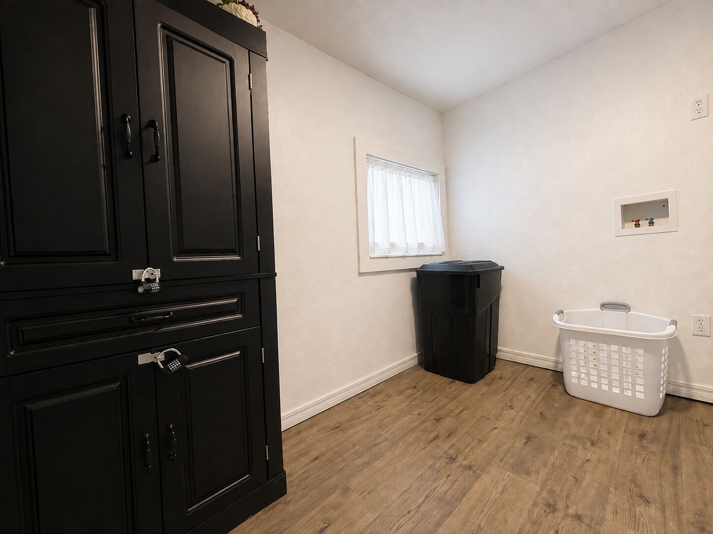 A room with a black cabinet, a trash bin, a laundry basket, and a window with white curtains.
