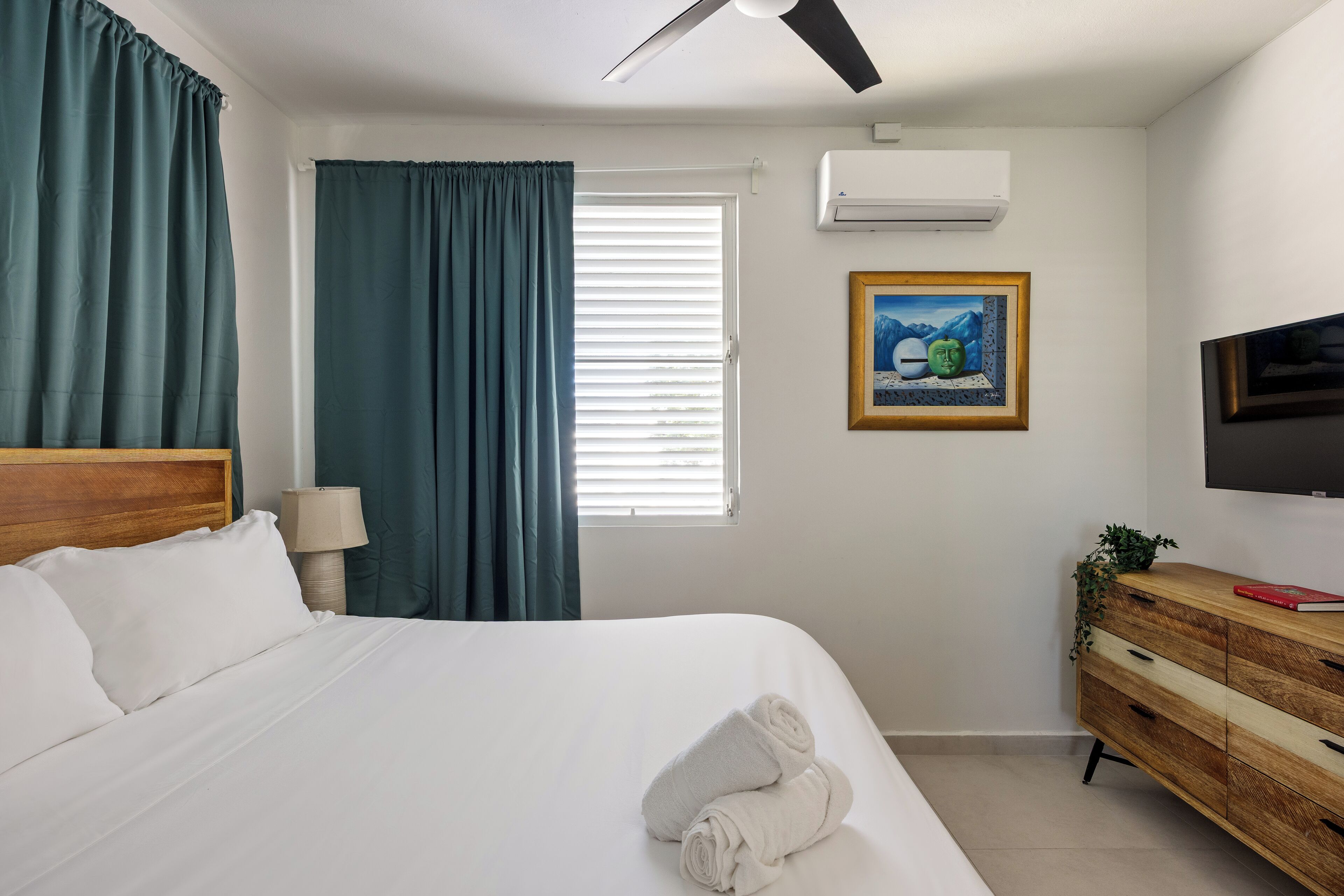 Quarto de hotel com uma cama, penteadeira de madeira, televisão, quadro e janela com cortinas.