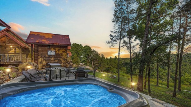Een hottub met een houten terras en buitenmeubilair, omgeven door een weelderig groen landschap en een houten chalet.