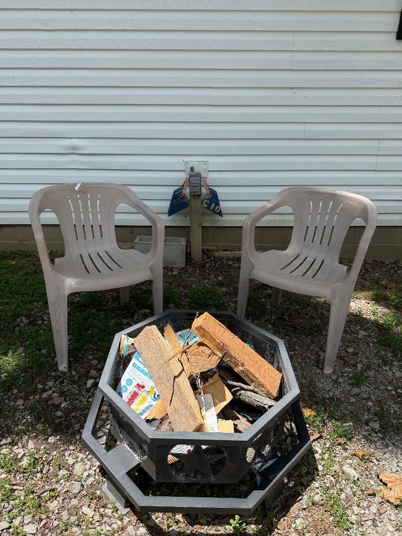 Two white plastic chairs, a hexagonal fire pit with wood and magazines, and a white building in the background.