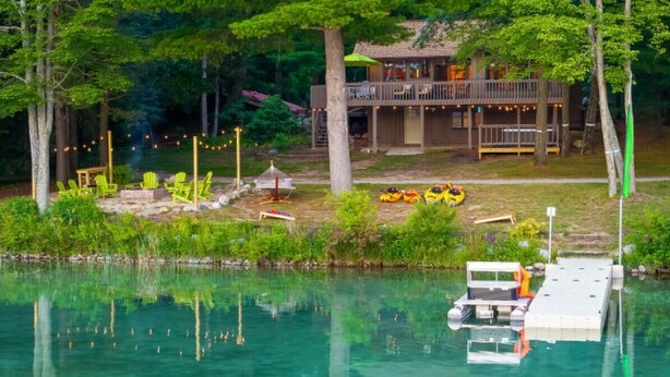 A lakeside cabin with a dock and kayaks, surrounded by trees and a clear view of the water. - Hot Tub, Water Amenities, Lakeshore Cottage (Newaygo)