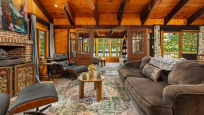 A cozy cabin interior with a fireplace, wooden ceiling beams, a large window, and a comfortable seating area. - Hot Tub, Water Amenities, Lakeshore Cottage (Newaygo)