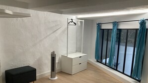 A modern bedroom with a white dresser, a black bench, a standing heater, and a large window with curtains.