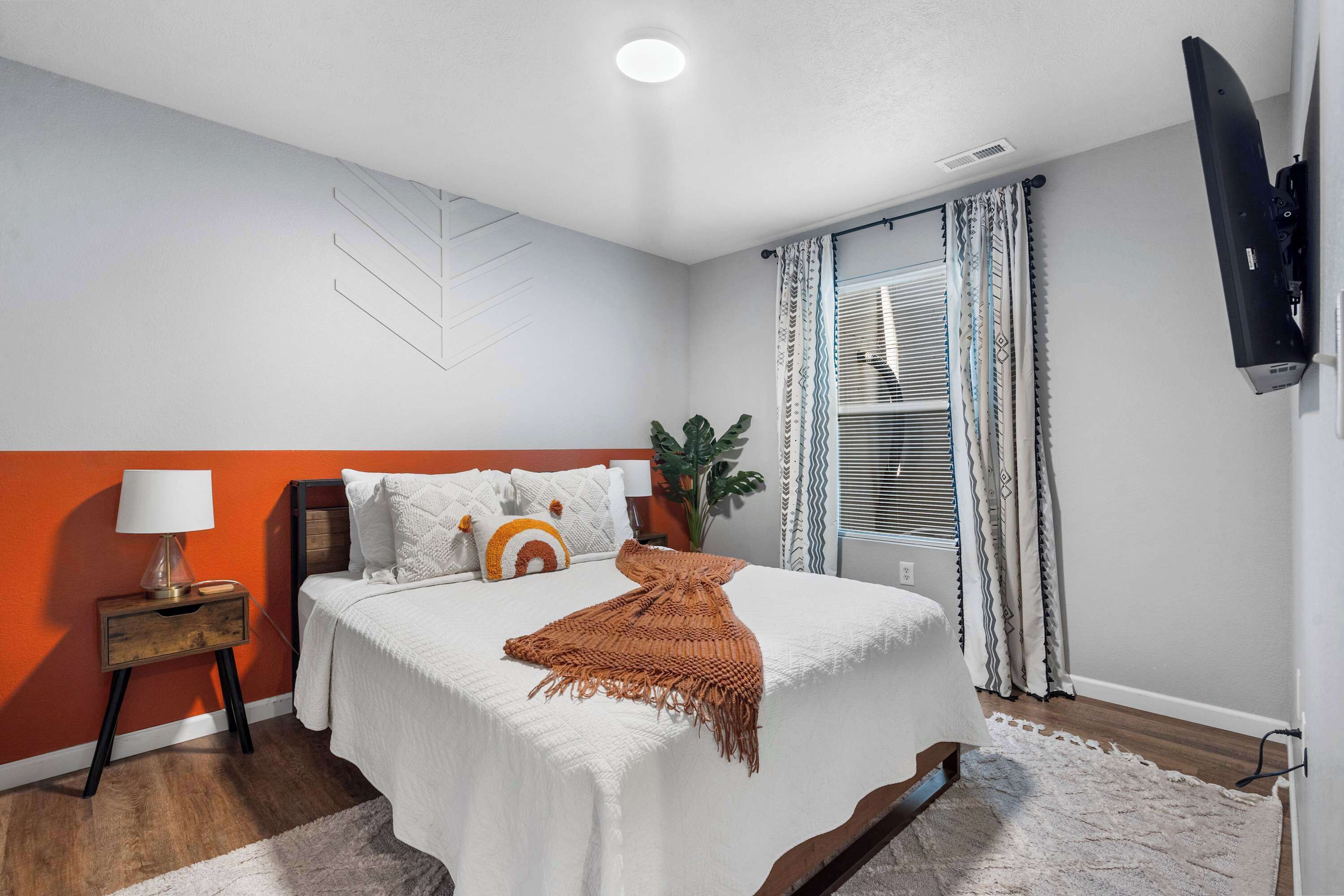 Un dormitorio con cama, mesita de noche, lámpara, televisor montado en la pared y ventana con cortinas.