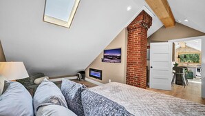 A bedroom with a sloped ceiling, a brick chimney, a TV, and a view of the kitchen.