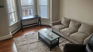 Ett vardagsrum med en beige soffa, ett soffbord i glas och ett svart sidobord. Det finns tre fönster med persienner, ett element och ett trägolv.