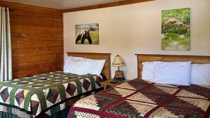 Una habitación con dos camas, paredes de madera, colcha acolchada, una lámpara y dos cuadros en la pared.