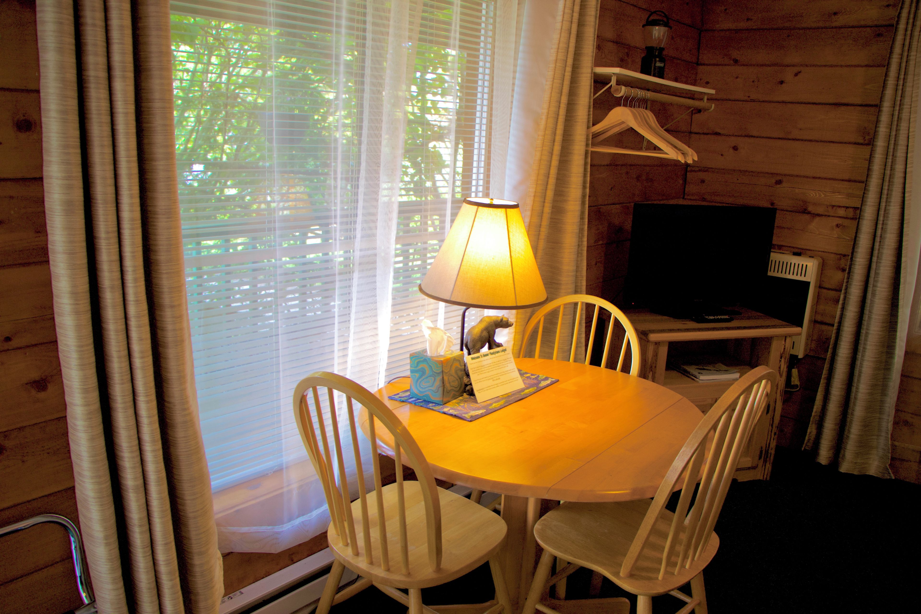 A wooden interior with a round table, two chairs, a lamp, a television, and a hanging coat rack.