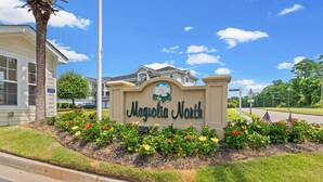 A sign for Magnolia North with a flower emblem, surrounded by a flower bed with various colors, a palm tree, and a building in the background. - Lucky Eagle @ Myrtlewood Golf (Myrtle Beach)