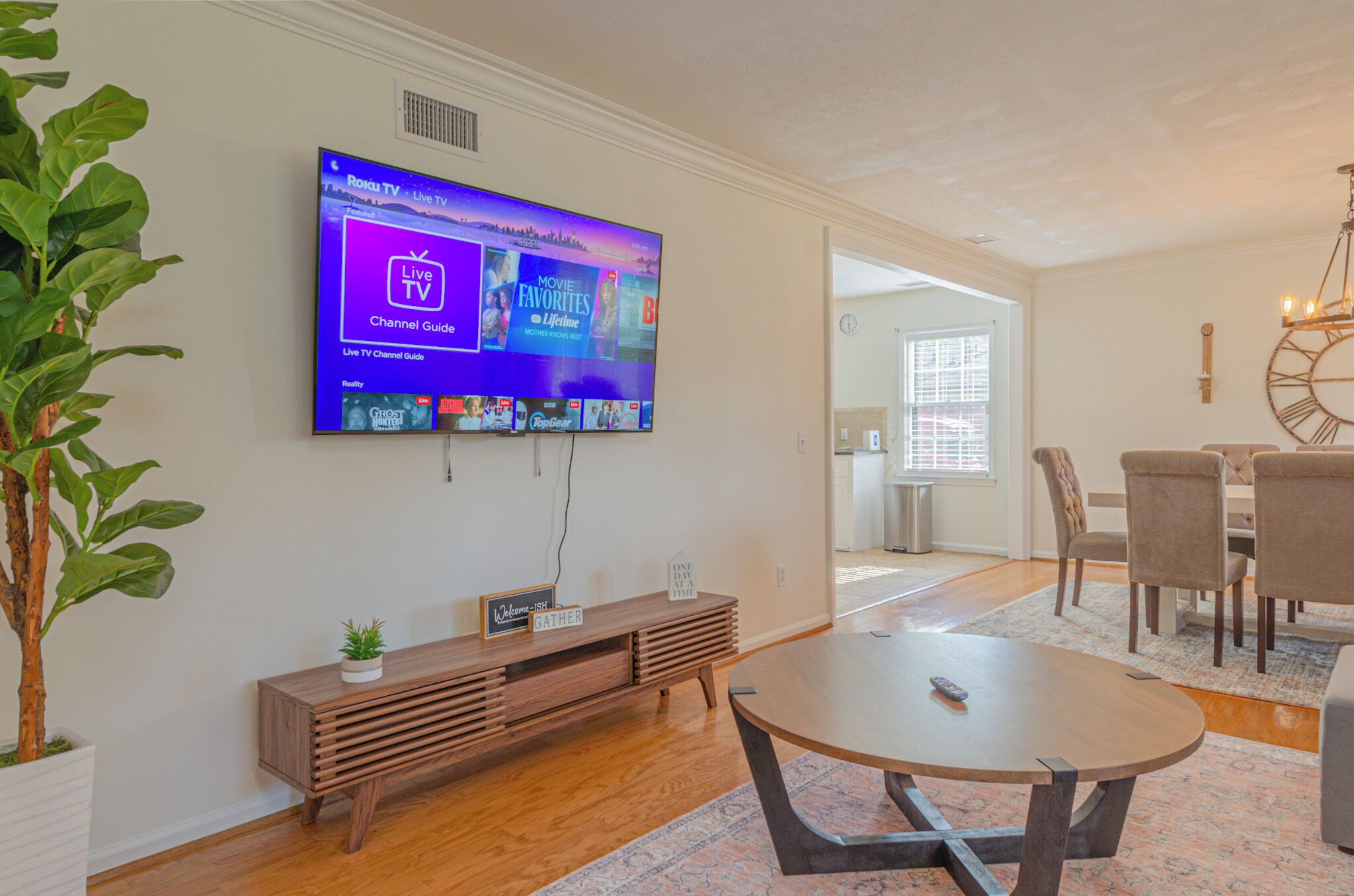Un salon moderne avec un téléviseur à écran plat fixé au mur, une console en bois, une table basse ronde et un coin repas avec des chaises et un lustre.