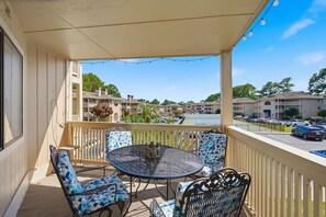 A balcony with a round table and chairs, a string of lights, and a view of a parking lot and buildings.