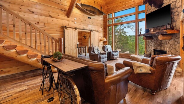 A rustic cabin interior with a wooden staircase, leather furniture, a stone fireplace, and a mounted deer head. - Buffalo Lodge - Stay for 8 (Branson)