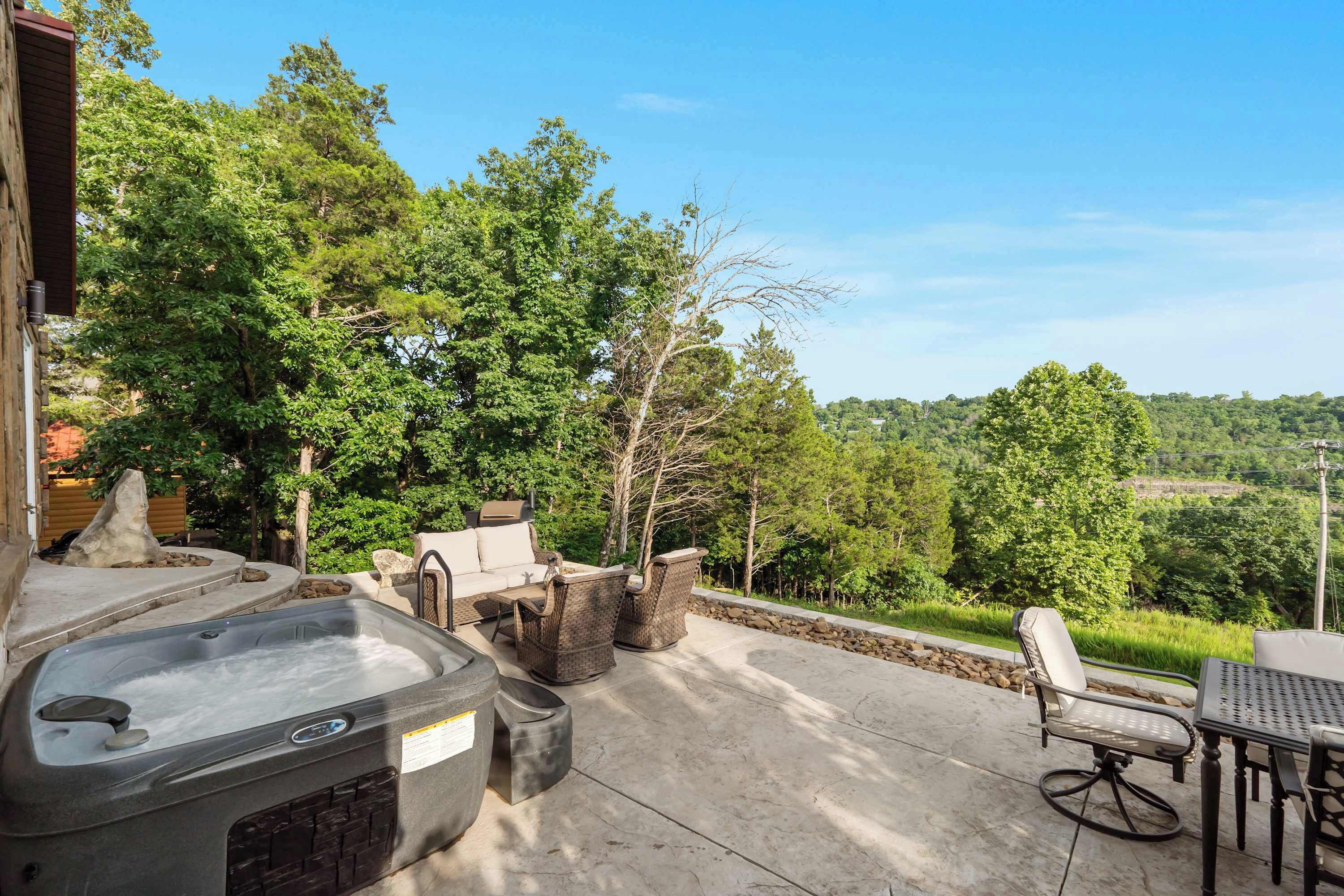 An outdoor patio with a hot tub, seating area, and a view of a forested landscape.