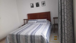 Ein Schlafzimmer mit einem Bett, einem Holz-Kopfteil, einem Nachttisch, einem Sessel und einem gemusterten Vorhang.