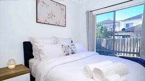 A neatly made bed with white linens and rolled towels, a bedside table with a lamp, and a window with a view of houses and a fence. - Family And Remote Work Friendly-Only 4 Minutes from The Pacific Werribee (Werribee)