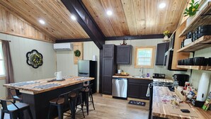 A kitchen with wooden flooring, a central island with stools, a black trash bin, and a window with a view. - The Barn in Kirby AR (Amity)