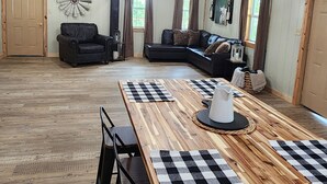 A spacious room with a wooden ceiling, a large wooden dining table with checkered placemats, two black leather sofas, and a chandelier. - The Barn in Kirby AR (Amity)