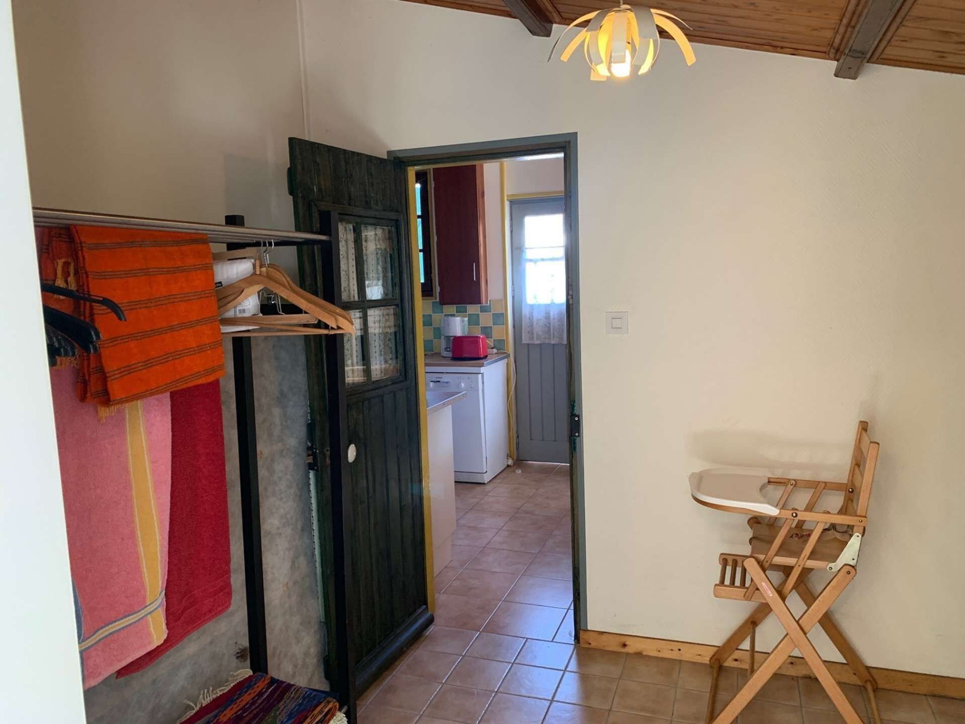 A room with a wooden floor, a folding chair, a hanging rack with clothes, and a kitchen visible through an open door.