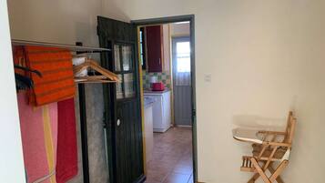 A room with a wooden floor, a folding chair, a hanging rack with clothes, and a kitchen visible through an open door.